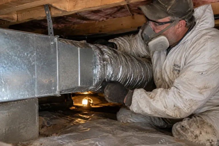 A technician wearing a dusty protective suit carefully replacing insulated ductwork in a crawl space beneath a home.