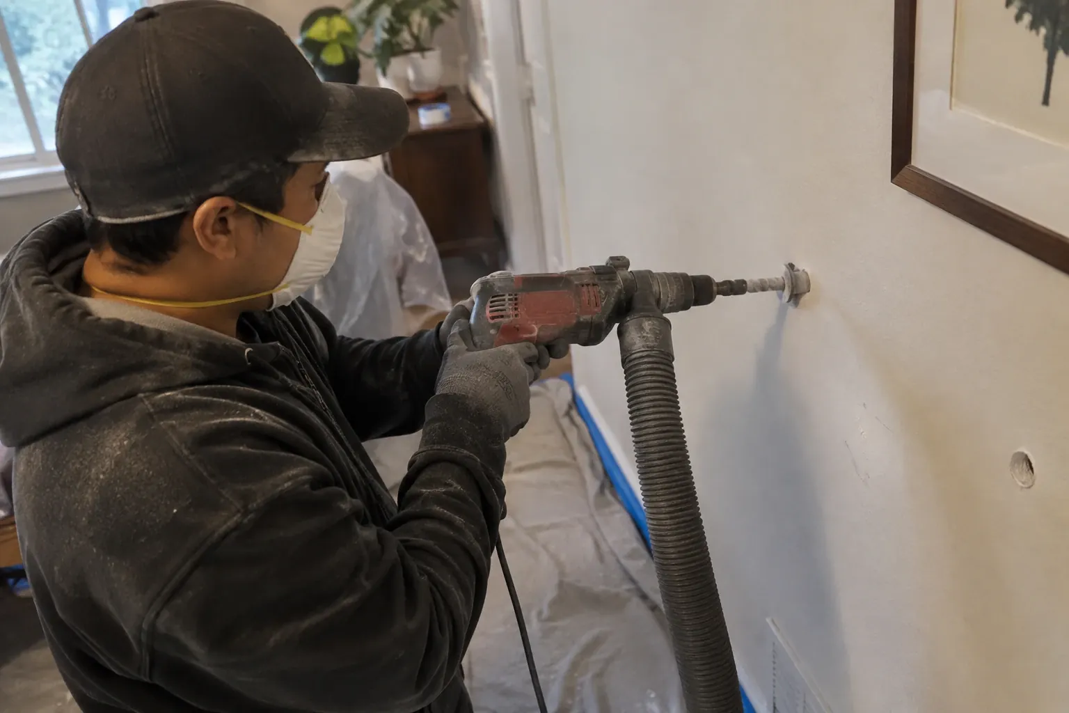 A technician performs cavity wall insulation using a drill-and-fill method inside a home, with floor protection clearly in place to prevent damage.