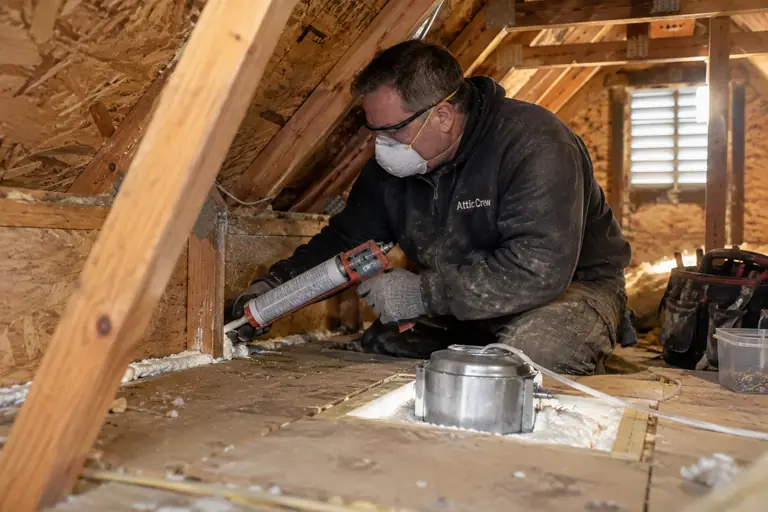 A technician is carefully air-sealing top plates and recessed lighting in an attic to prepare for insulation installation.