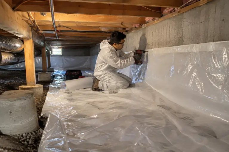 A technician is fastening and sealing a heavy white moisture barrier liner securely to the concrete foundation wall of a crawl space.