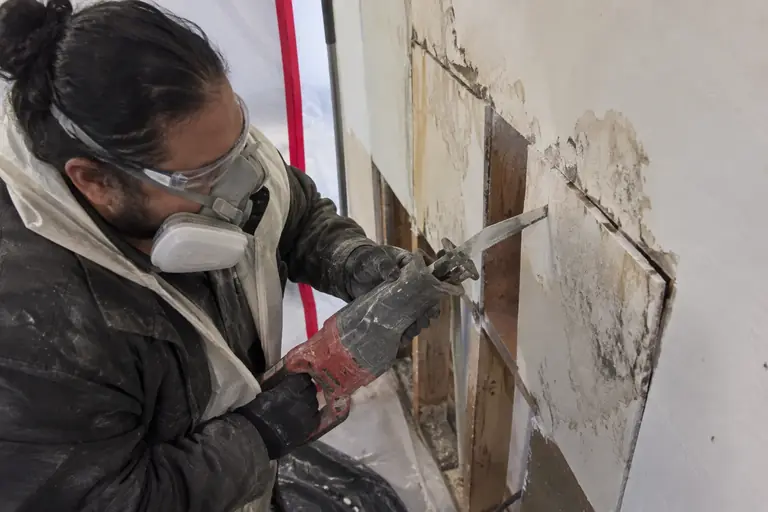 A technician carefully cuts out sections of water-damaged drywall inside a sealed containment area during mold remediation.