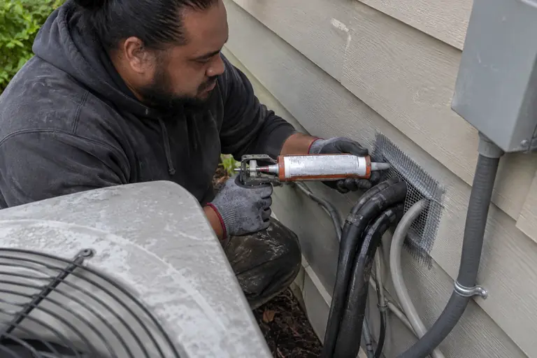 A technician is sealing a gap around AC lines and cable entry on the exterior of a home using heavy-gauge steel mesh and sealant.